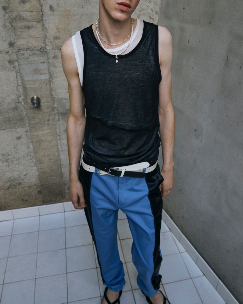 Young adult poses in a black tank top and blue and black color block pants, standing against a concrete wall and tiled floor.