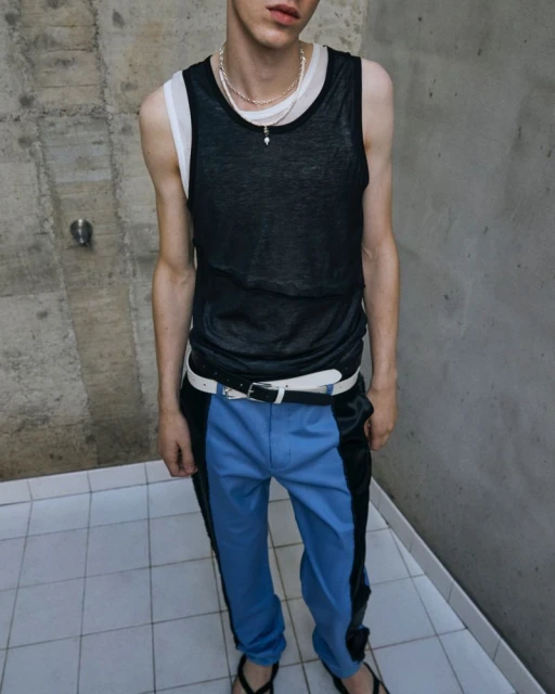 Young adult poses in a black tank top and blue and black color block pants, standing against a concrete wall and tiled floor.
