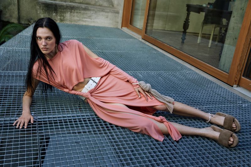 Woman in pink dress poses on metal grate outdoors. She is lying down looking at the camera wearing brown sandals and a white belt.