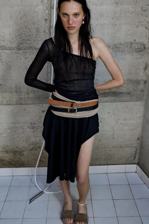 Model in dark sheer one-shoulder top and layered belts standing against textured concrete wall