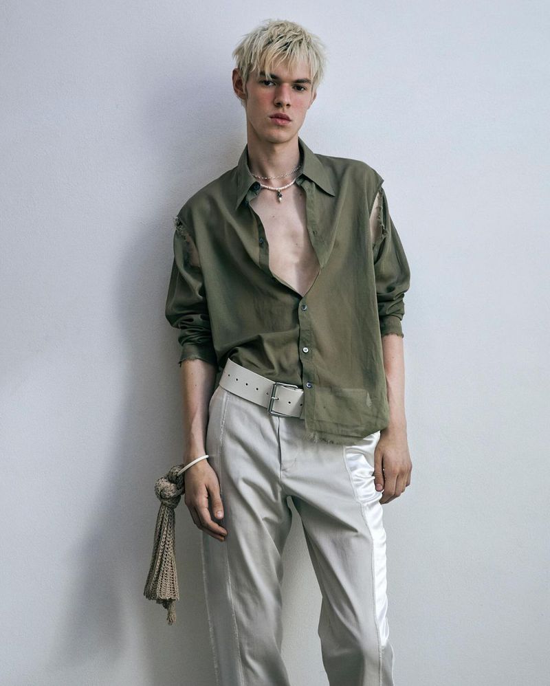 Blonde male model wearing an olive green shirt and white trousers standing against a plain white studio background.