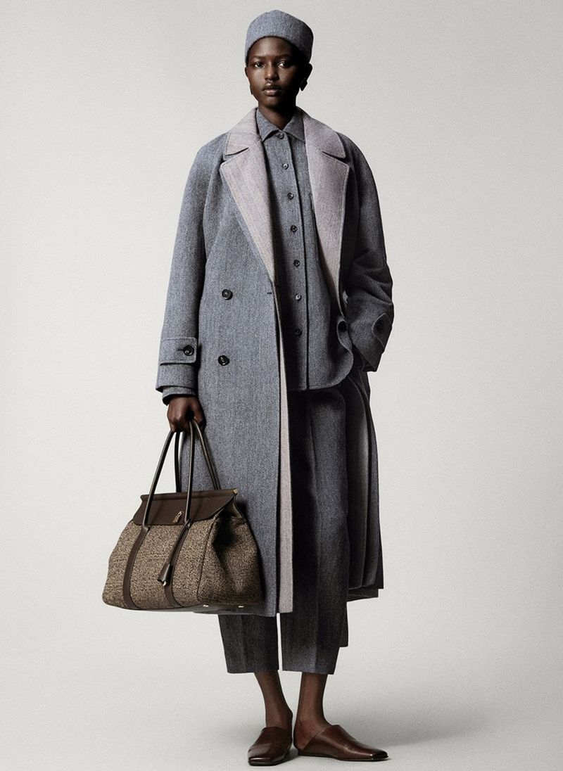 Elegant Black woman in herringbone grey wool overcoat, matching pants, shirt, and hat, carrying a brown leather bag, studio shot.