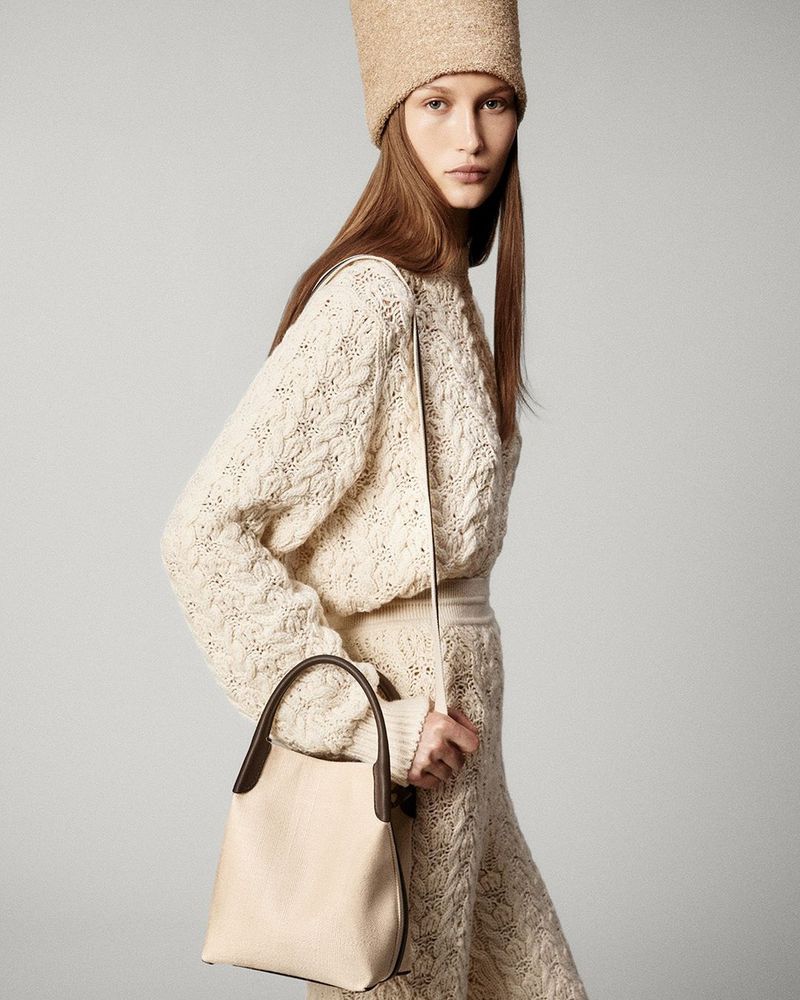 Blonde woman in knitted outfit and hat poses with a cream-colored handbag against a neutral studio backdrop in a fashion editorial.