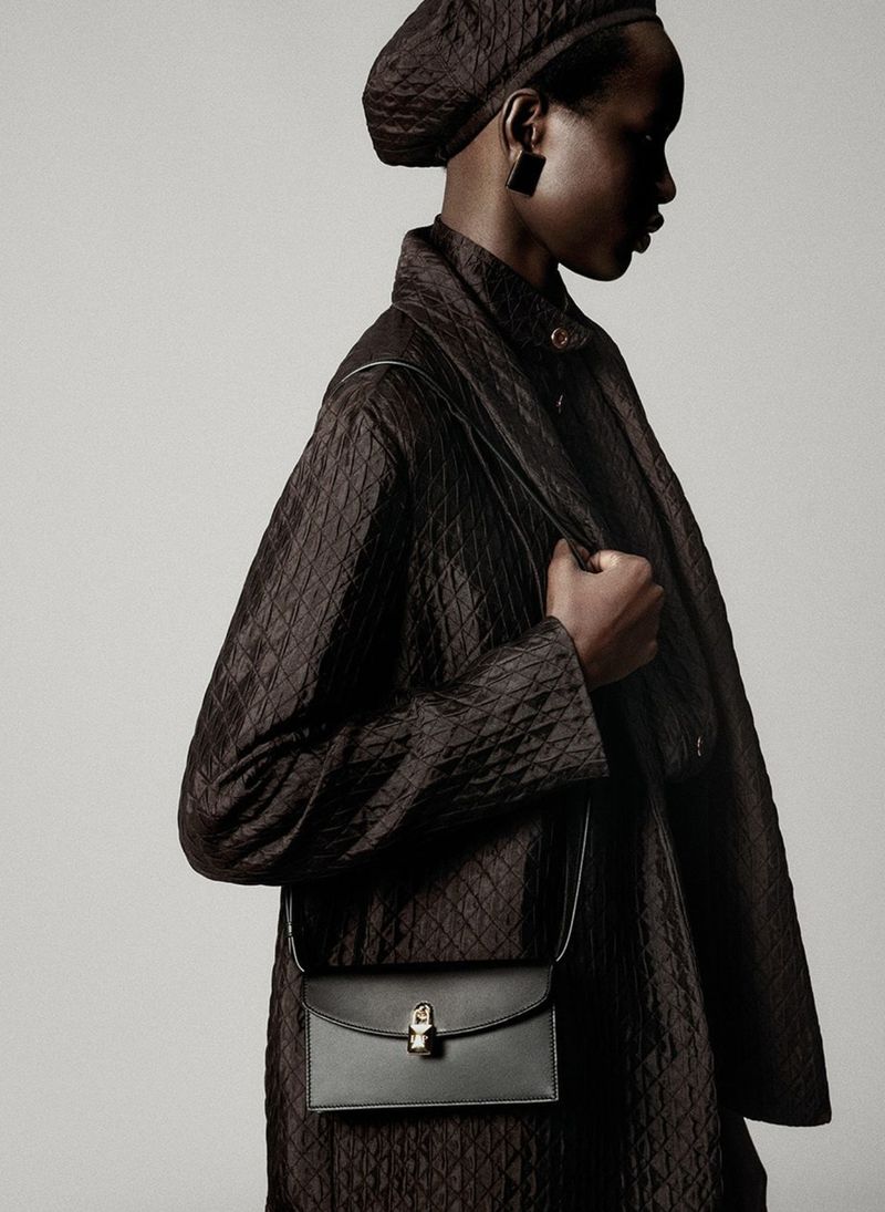 Profile shot of a Black female model in a quilted brown coat, matching hat and black purse on a light grey studio backdrop.