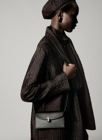 Profile shot of a Black female model in a quilted brown coat, matching hat and black purse on a light grey studio backdrop.