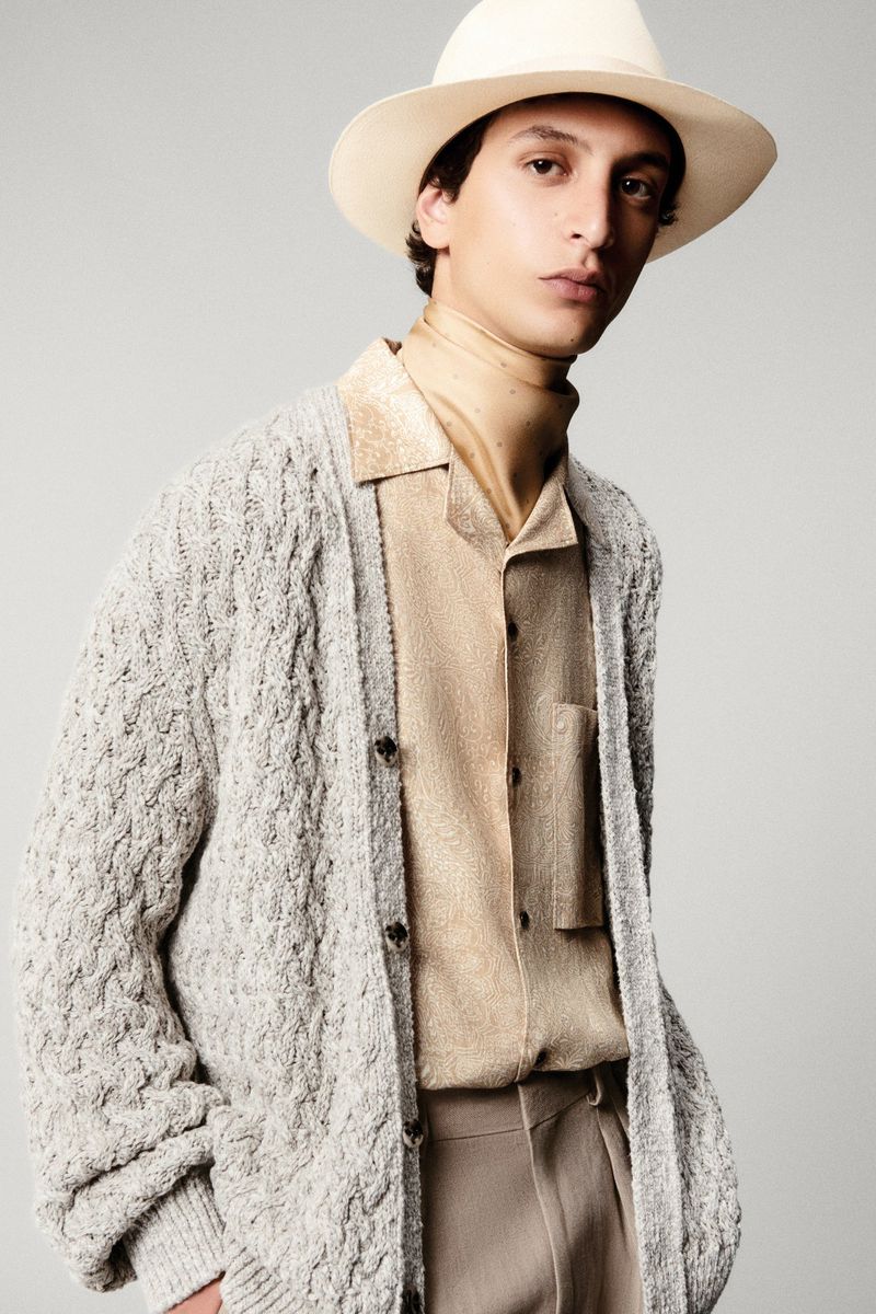 Portrait of a fashionable adult male wearing a light gray knit cardigan, a beige button-down shirt, beige pants, and a beige hat.