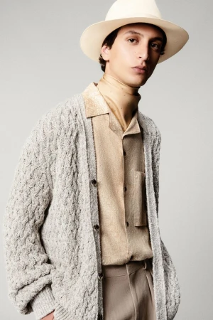 Portrait of a fashionable adult male wearing a light gray knit cardigan, a beige button-down shirt, beige pants, and a beige hat.