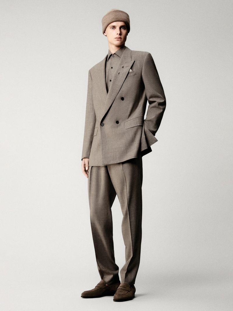 Male fashion model wearing a tan-colored suit and beanie, posing in a photo studio against a grey backdrop, looking directly at the camera.