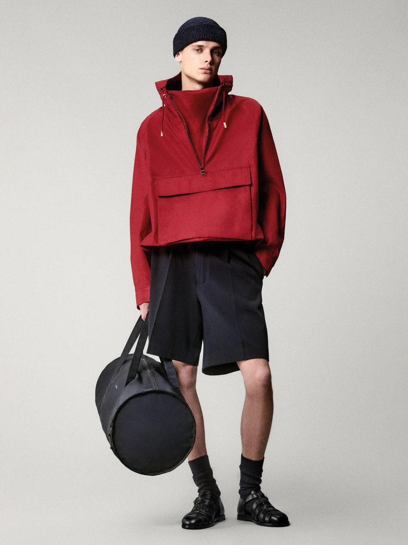 Male fashion model poses in a red zip-up jacket, black shorts, shoes, socks, and a beanie while holding a black duffel bag.