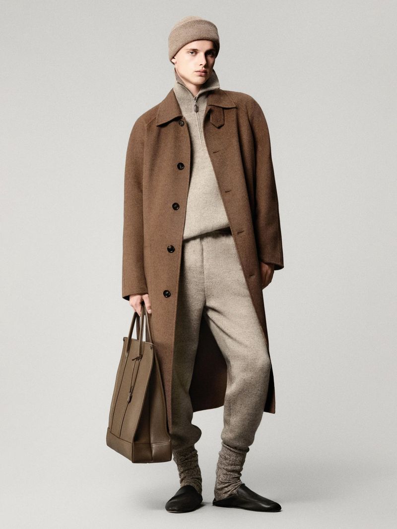 Man in brown overcoat, beige knitwear, sweatpants, hat, leather tote bag, and slippers posing in a studio against a grey backdrop.
