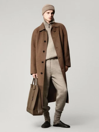 Man in brown overcoat, beige knitwear, sweatpants, hat, leather tote bag, and slippers posing in a studio against a grey backdrop.