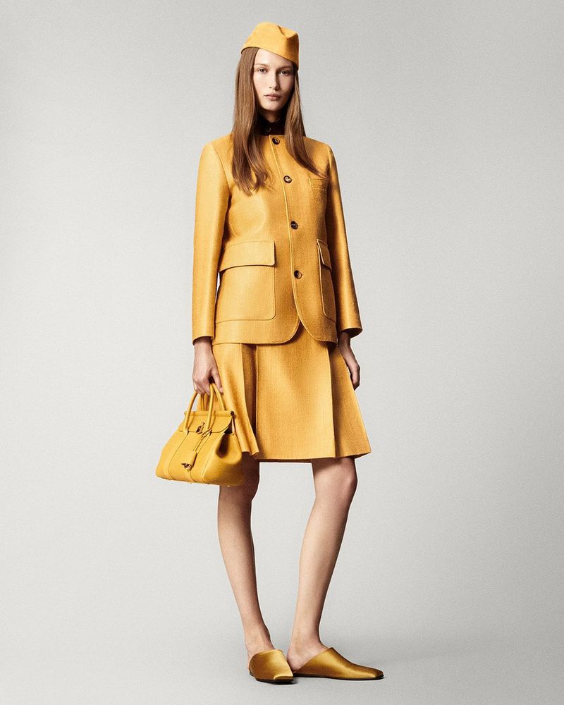 A woman models mustard-yellow apparel consisting of a skirt, jacket, hat, and handbag, complemented by flat shoes against a gray backdrop.