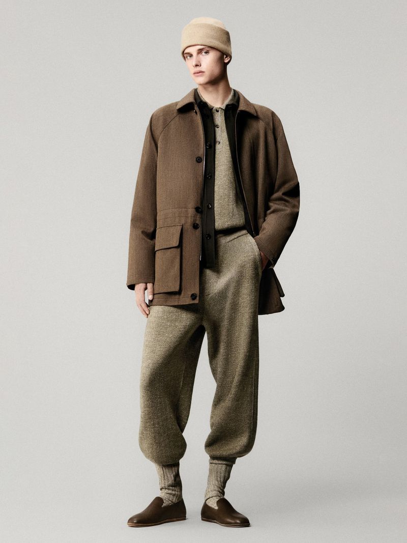 Adult male model in brown coat, beige knit sweater, and knit pants, stands indoors against a neutral background in studio lighting.