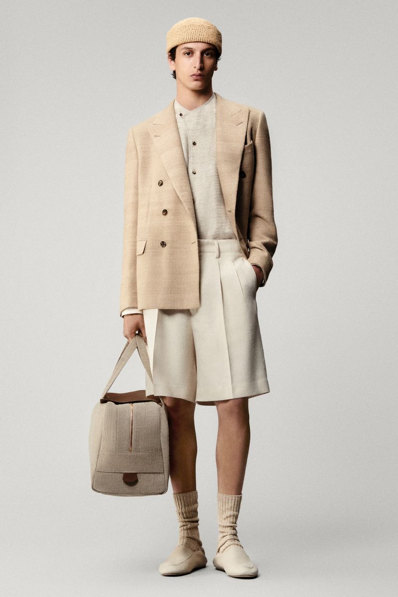 Male fashion model in a beige suit, shorts, and hat stands with a travel bag against a plain background in a studio shot.