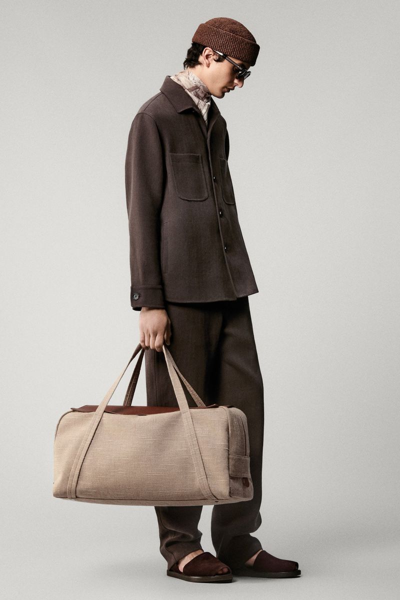 Man in a brown outfit and beanie carrying a beige duffel bag against a neutral background, showcasing a blend of fashion and utility.