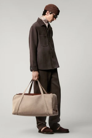 Man in a brown outfit and beanie carrying a beige duffel bag against a neutral background, showcasing a blend of fashion and utility.