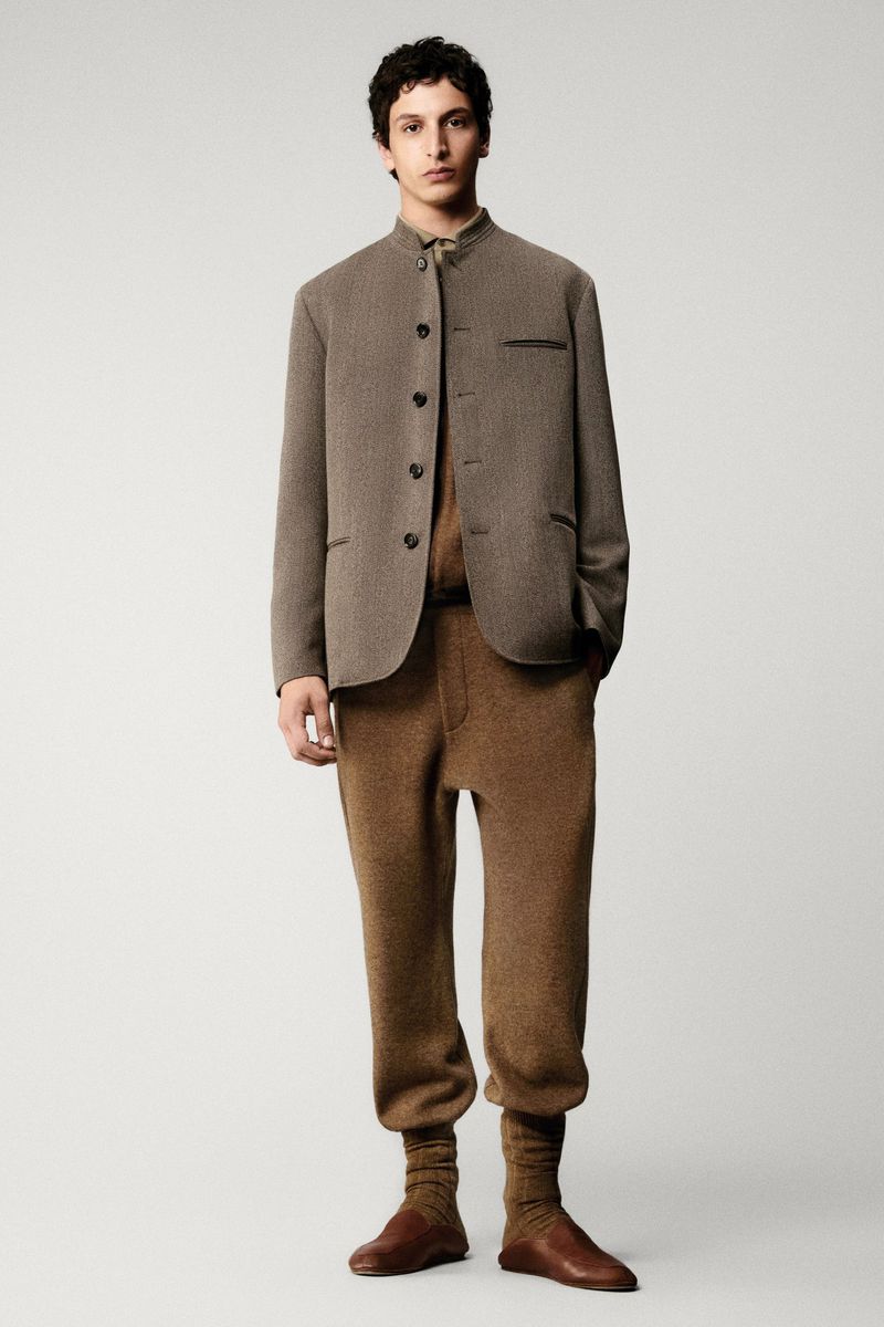 A young adult male is posing in a studio wearing a brown jacket, matching brown trousers, and brown leather shoes, indoor on a neutral background.