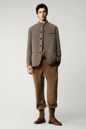 A young adult male is posing in a studio wearing a brown jacket, matching brown trousers, and brown leather shoes, indoor on a neutral background.