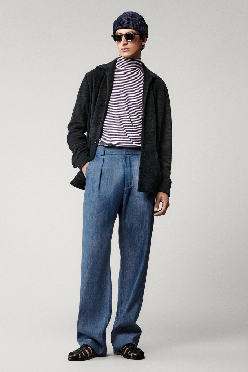 Man posing in striped turtleneck, blue jeans, black jacket, beanie, and sunglasses against a neutral backdrop in a studio shot.