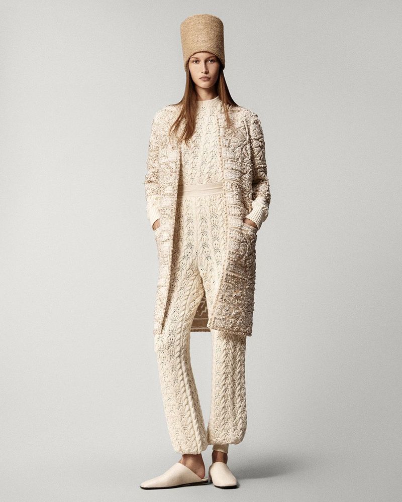 Woman in a cream-colored, textured knit outfit, including a hat, sweater, coat, and pants, standing against a neutral backdrop in a studio setting.