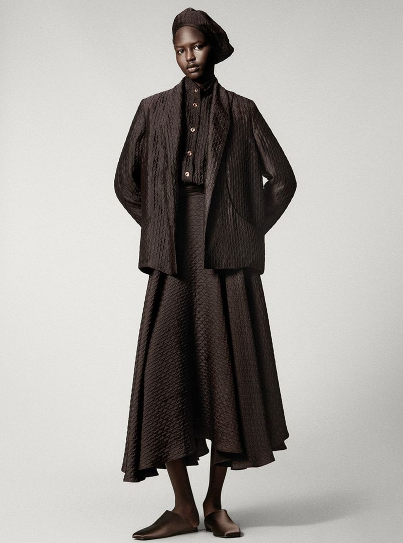 A full-length shot of an adult female fashion model in brown patterned, traditional-style clothing against a neutral background in a studio.