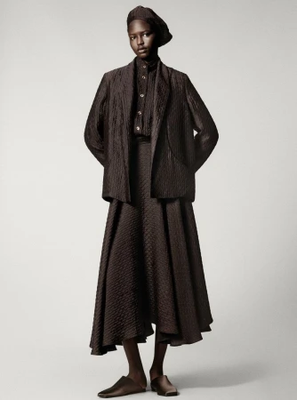 A full-length shot of an adult female fashion model in brown patterned, traditional-style clothing against a neutral background in a studio.