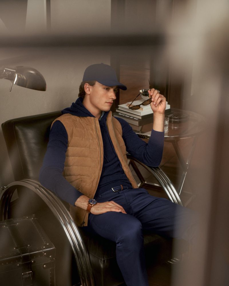 A young man wearing a vest, baseball cap, and sweater sits in a chair holding his sunglasses inside a home with neutral colors.