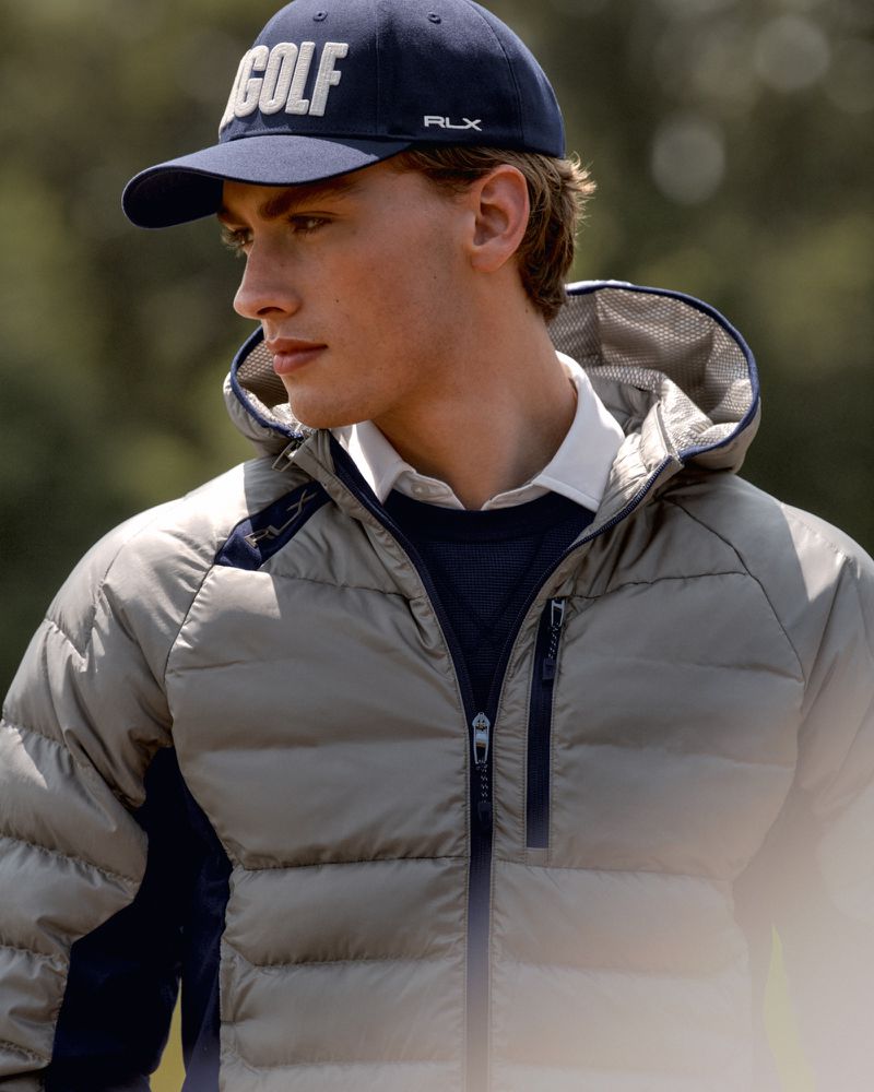 A young adult male wearing a navy baseball cap with the word 'GOLF' and a puffer jacket poses in a close-up, outdoor shot.