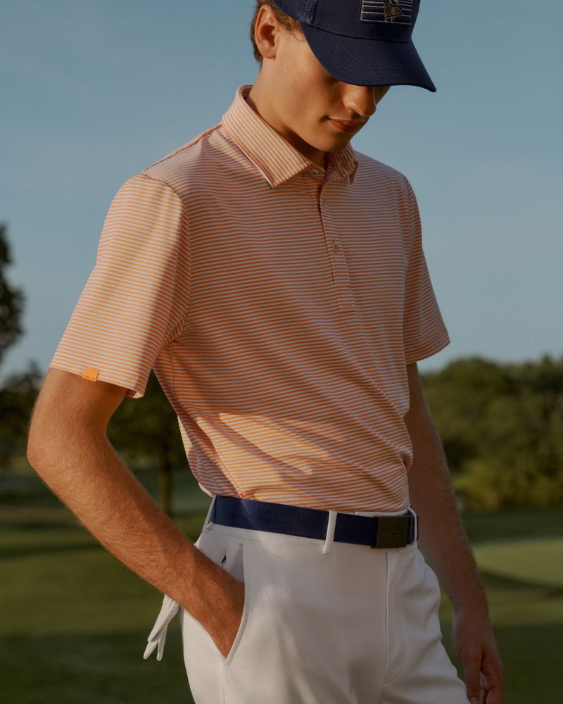 A person wears a horizontally striped peach and white polo shirt, white pants, and a navy baseball cap in an outdoor setting.