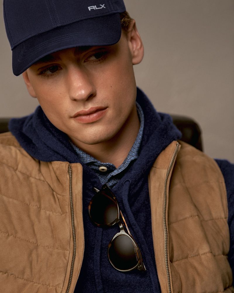 Portrait of a young man wearing a navy baseball cap, vest, and sweater, looking to the side with neutral expression in a studio setting.