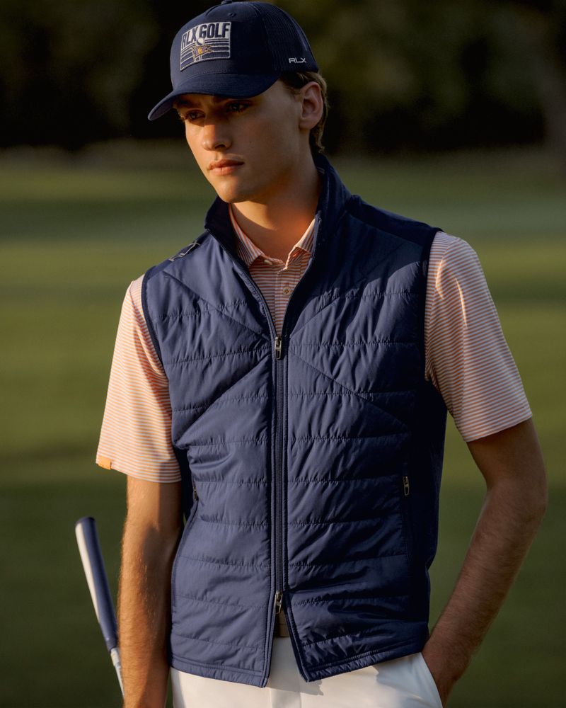 A fair-skinned young man in a sleeveless puffer vest and RLX Golf cap stands on a green golf course in a medium shot.