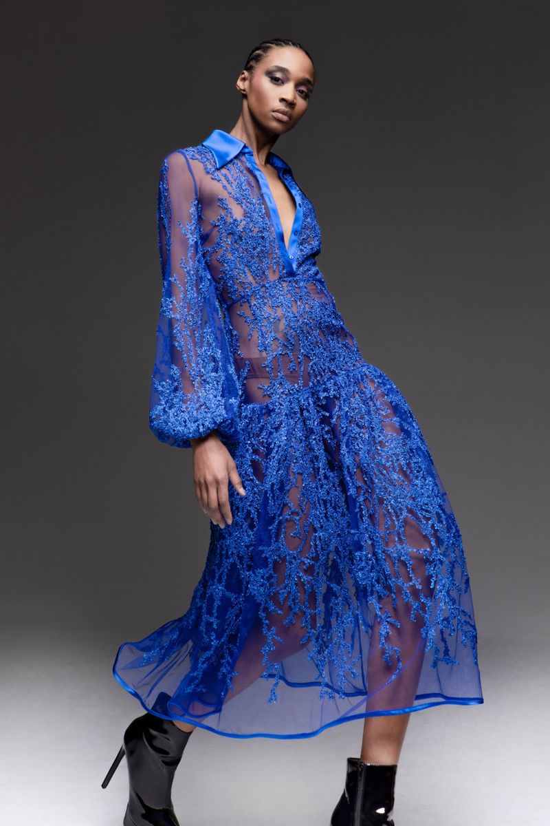 Fashion model posing in a sheer blue dress with long sleeves and black boots, set against a neutral gray studio backdrop. 