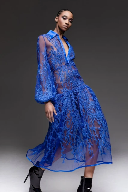 Fashion model posing in a sheer blue dress with long sleeves and black boots, set against a neutral gray studio backdrop. 