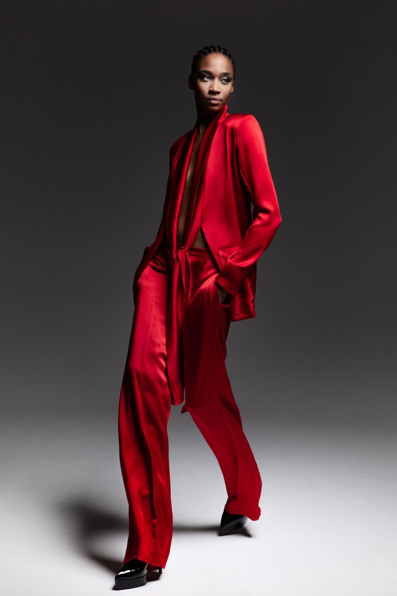 A person with cornrows in red satin suit stands in a photo studio, looking off-camera with a serious expression.