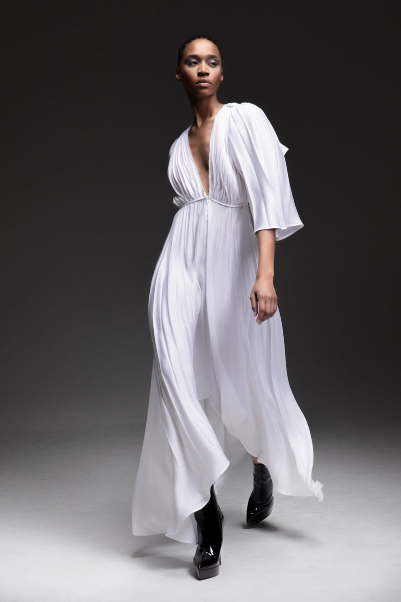 Woman in flowing white gown and black patent leather boots poses in a studio with a dark background, fashion editorial style.