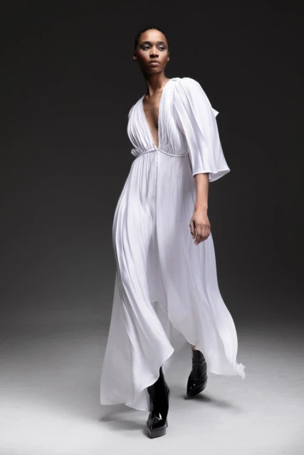Woman in flowing white gown and black patent leather boots poses in a studio with a dark background, fashion editorial style.