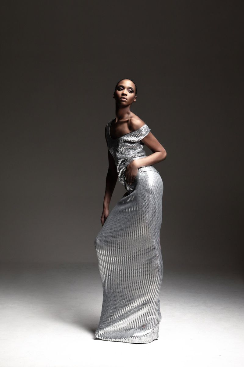 Elegant woman posing in a shimmering silver evening gown in a studio setting, showcasing fashion and style with a dramatic spotlight effect.