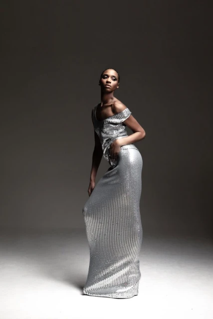 Elegant woman posing in a shimmering silver evening gown in a studio setting, showcasing fashion and style with a dramatic spotlight effect.