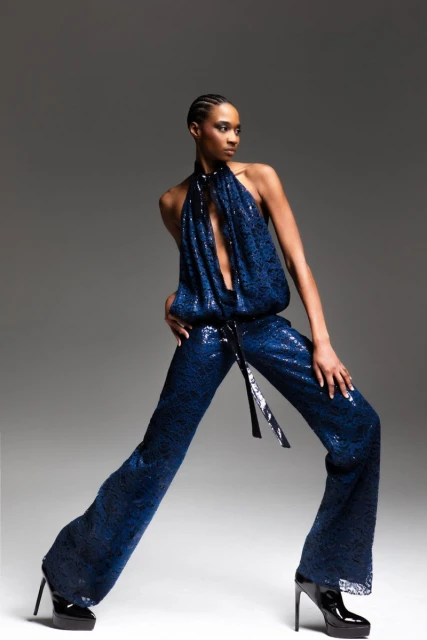 A Black female model poses in a blue sequined halter jumpsuit with wide legs and black high heels in a fashion photo shoot.
