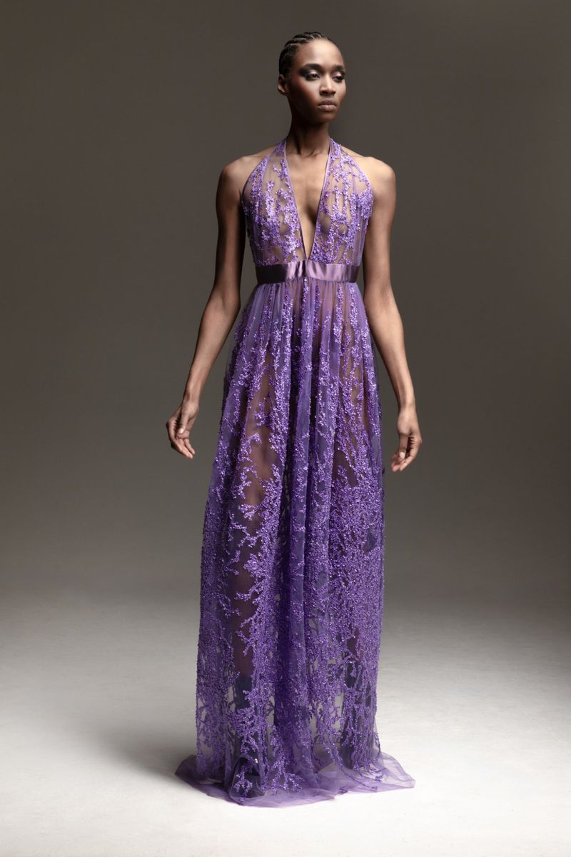 Full-length portrait of a Black female fashion model posing in a floor-length, semi-sheer purple gown in a photo studio setting.