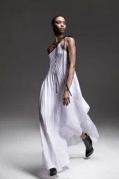 Model in a white pleated dress with black straps walking against a grey seamless background
