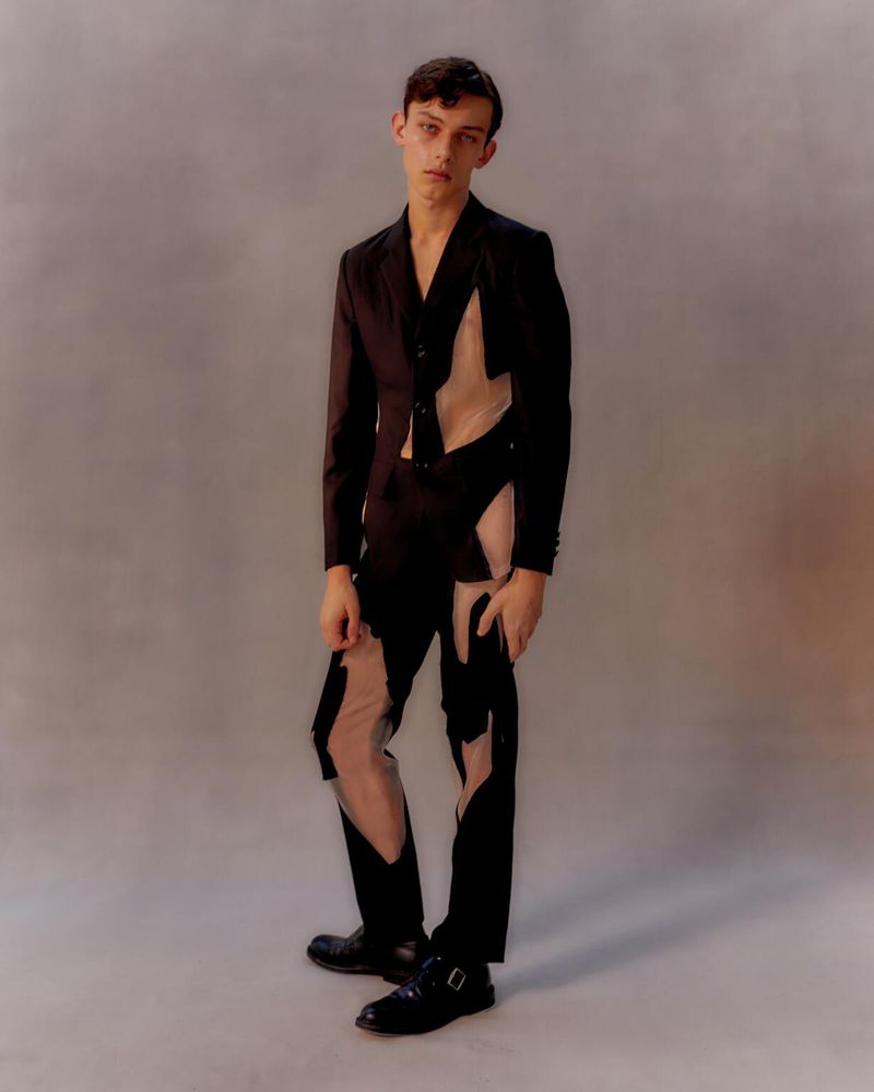 Full-length portrait of a young adult male in a black avant-garde deconstructed suit, posing in a photo studio setting on a light gray background.