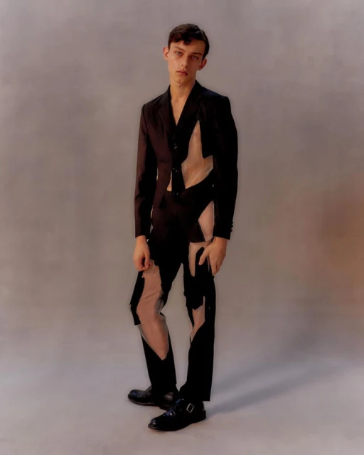 Full-length portrait of a young adult male in a black avant-garde deconstructed suit, posing in a photo studio setting on a light gray background.