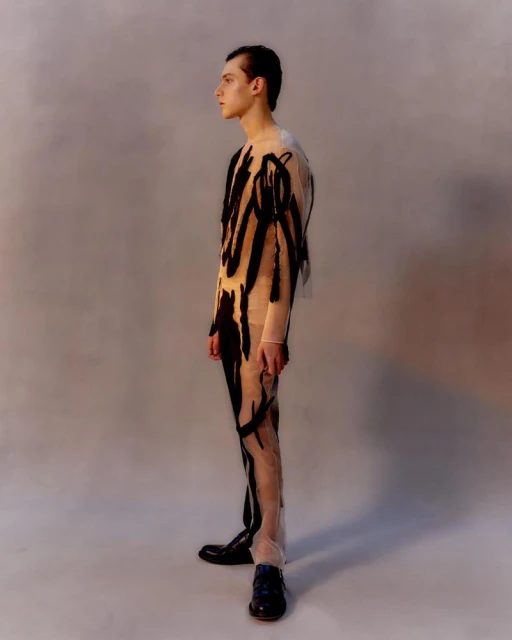 Studio shot of a young adult male fashion model wearing a sheer bodysuit with black abstract pattern, looking off to the side in a studio.