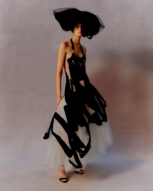 A woman in a black and white dress and oversized black hat poses in a studio setting, showcasing a dramatic fashion ensemble.