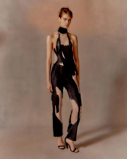 Full-length shot of a model in a black, halter-neck jumpsuit with open cutouts, wearing high heels, posing in a studio with soft lighting.