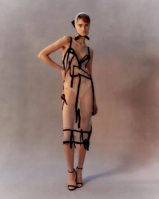 Full-length shot of a woman in a sheer black-trimmed dress and headscarf, posing in black heels against a muted studio backdrop.