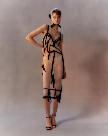 Full-length shot of a woman in a sheer black-trimmed dress and headscarf, posing in black heels against a muted studio backdrop.