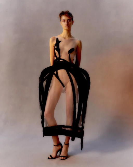 Fashion photo of a woman in a sheer dress with painted details, posing in high heels against a plain studio backdrop.