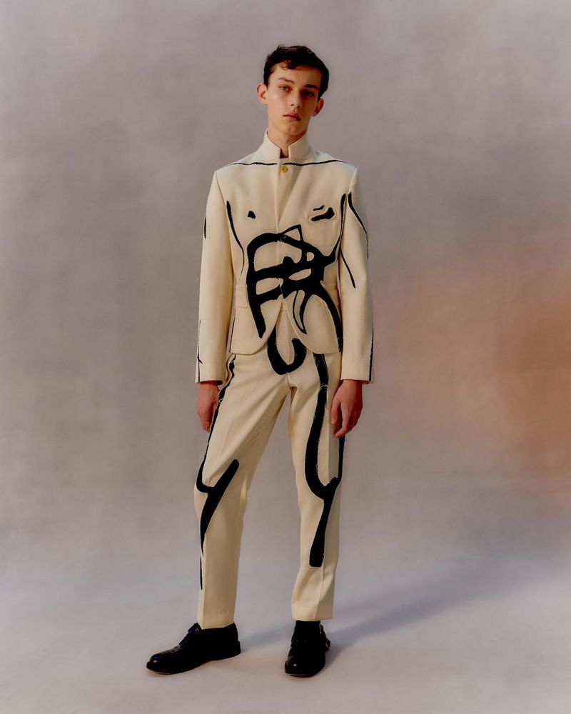 Adult male model posing in a studio wearing a cream-colored pantsuit with black abstract graphic art, and black patent leather shoes.